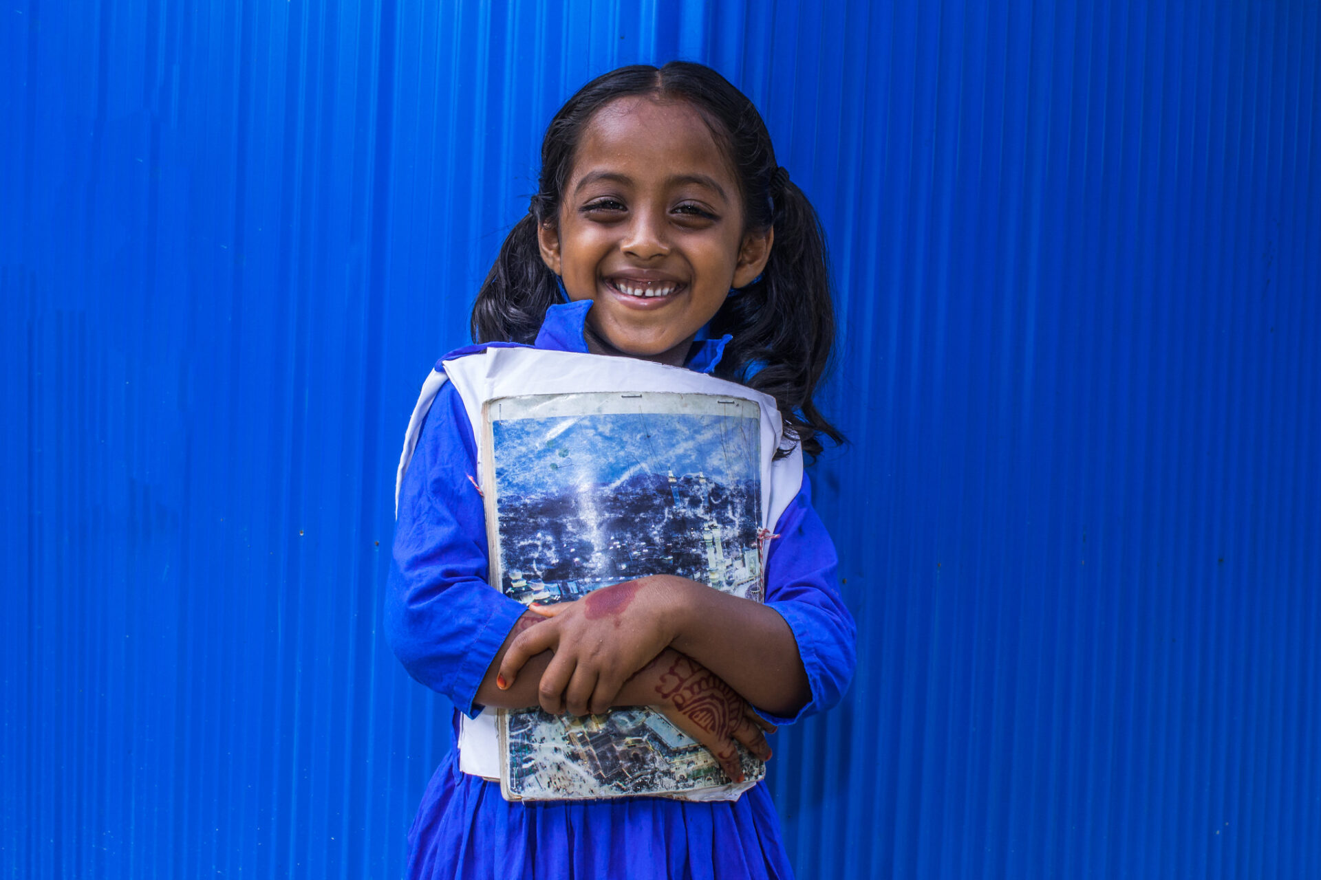 A young girl, with a brilliant smile, stands in front a bright blue background that matches her new school dress.