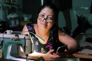A female tailor with glasses holding a phone in front of a sewing machine in Vietnam