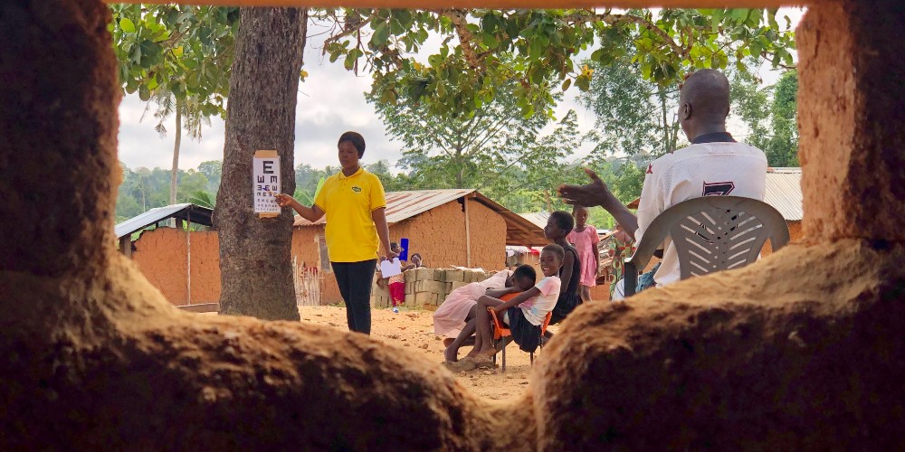 Image description: A photograph is taken through the window of a mud house. It frames a vision screening in progress where an eye care professional points at a snell chart. 