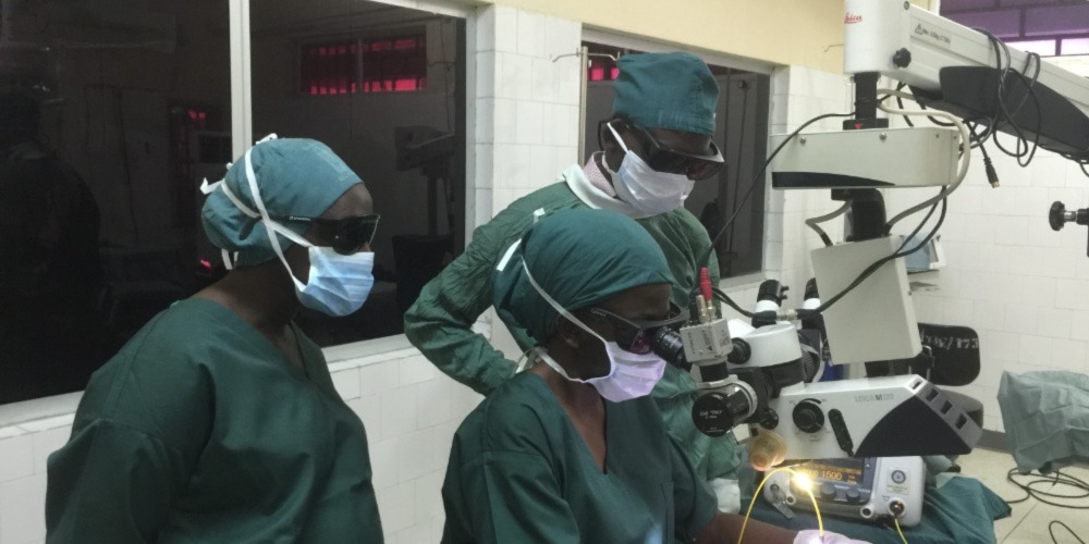 3 people wearing green scrubs and masks peer into a microscope