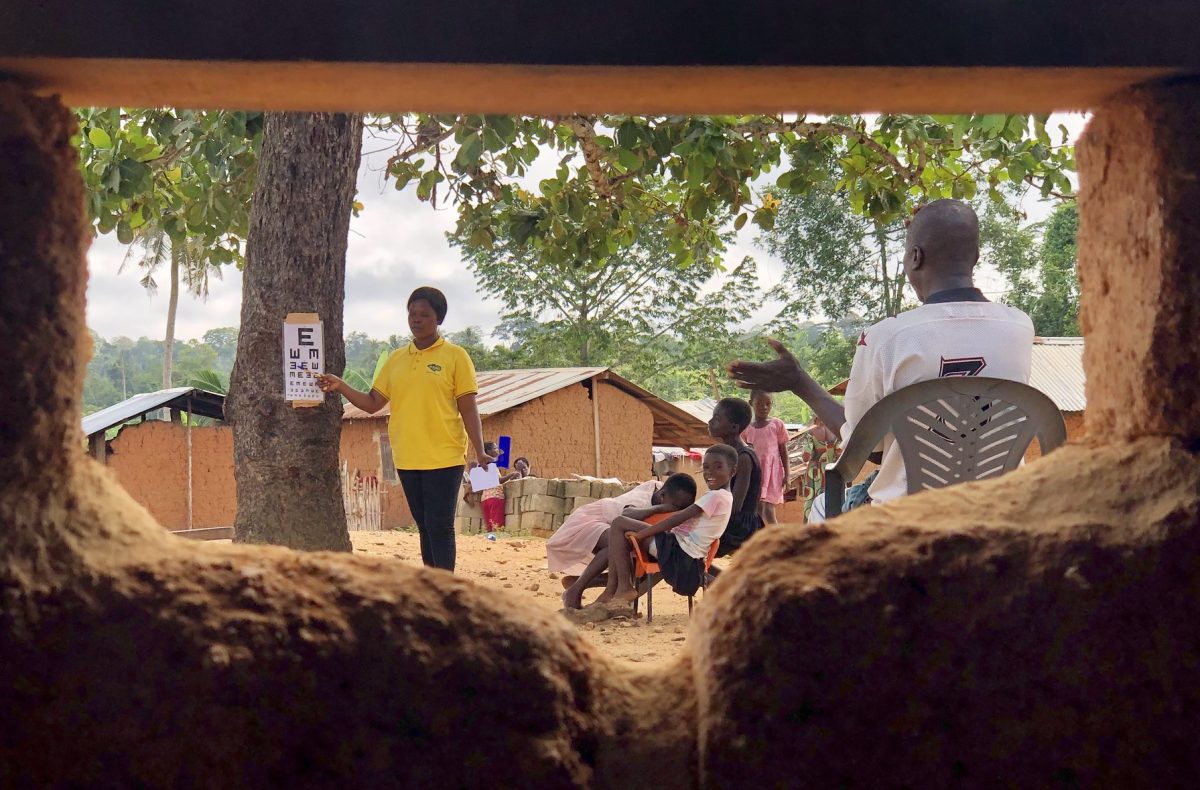 Image description: A photograph is taken through the window of a mud house. It frames a vision screening in progress where an eye care professional points at a snell chart. 