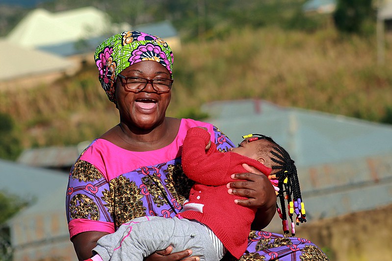 An african woman laughing in full traditional dress holding her child in her arms - wearing glasses