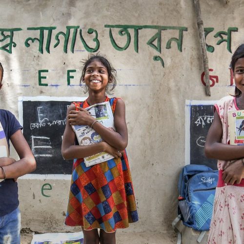 Three young girls in a classroom, smiling and holding books, stand in front of a wall with text and drawings. They appear happy and engaged, showcasing their books.