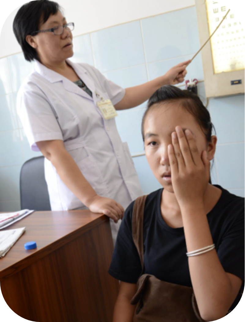 A healthcare worker in a white coat points to a vision chart while a woman covers one eye during an eye test.