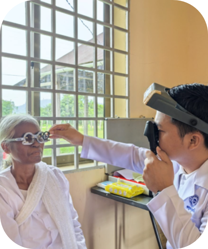 A male healthcare worker examines the eyes of an older woman wearing a trial frame in a clinic with barred windows.