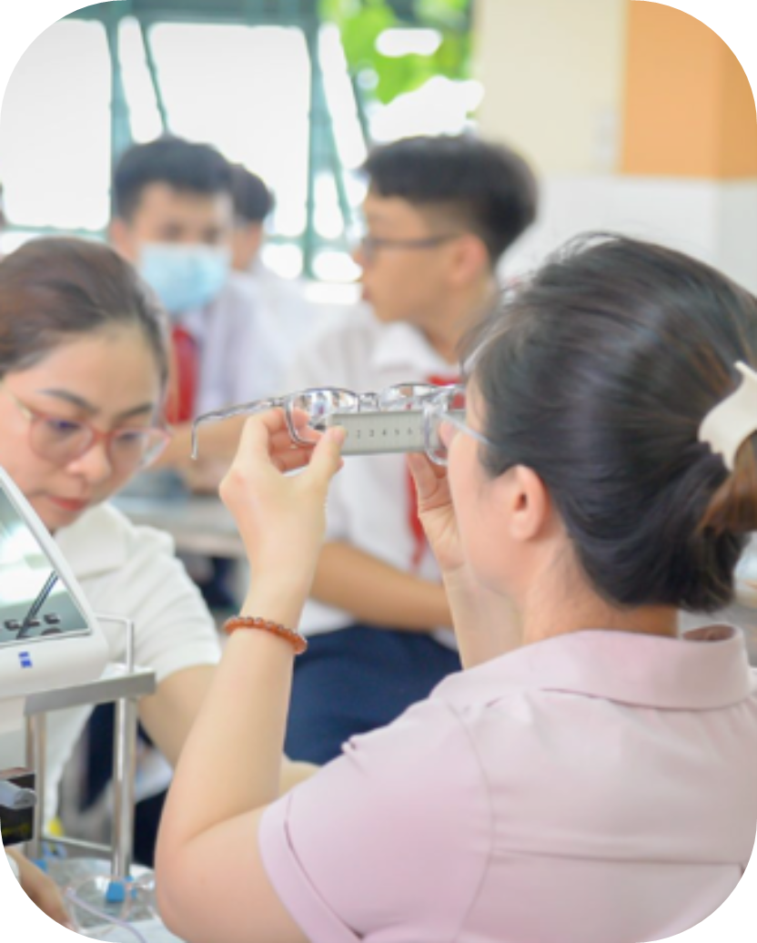 A woman inspects a pair of glasses using a ruler, with students in uniform blurred in the background.
