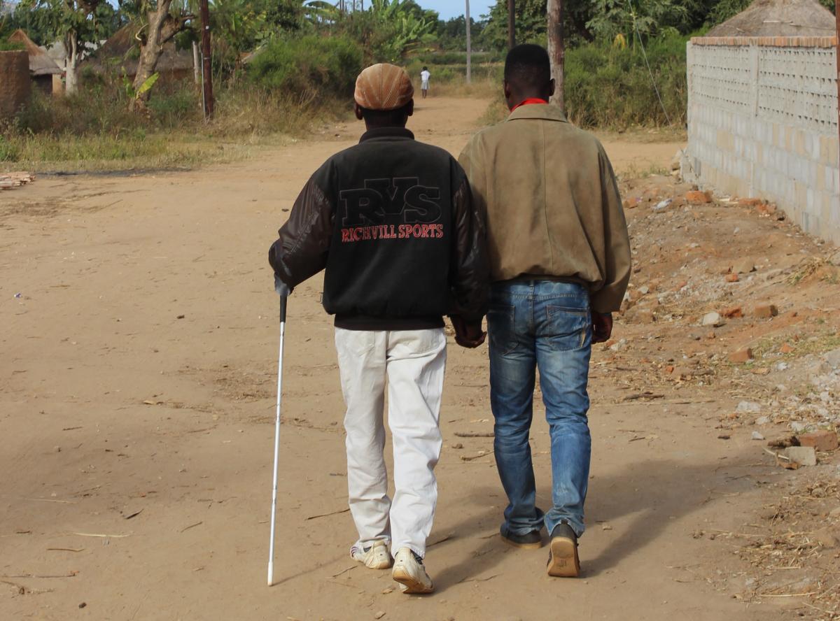 A blind man using a white cane, being assisted by another man