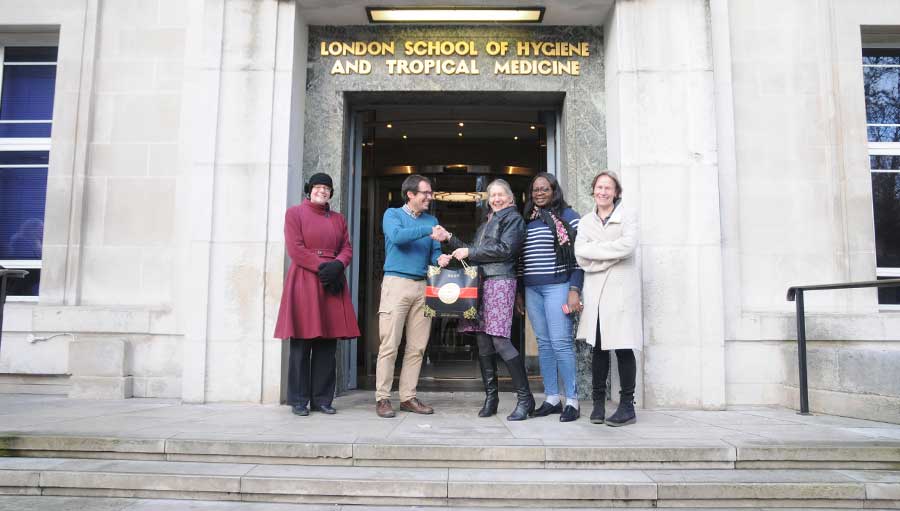 The winner is standing at the entrance of the London School of Hygiene and Tropical Medicine building with Sally Crook who is handing over a bag containing a DSLR camera. Three IAPB team members standing on each side are smiling at the camera